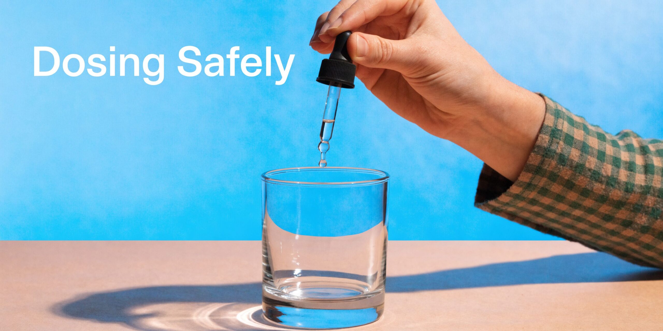 A person holding a glass dropper over an empty clear drinking glass against a blue background.