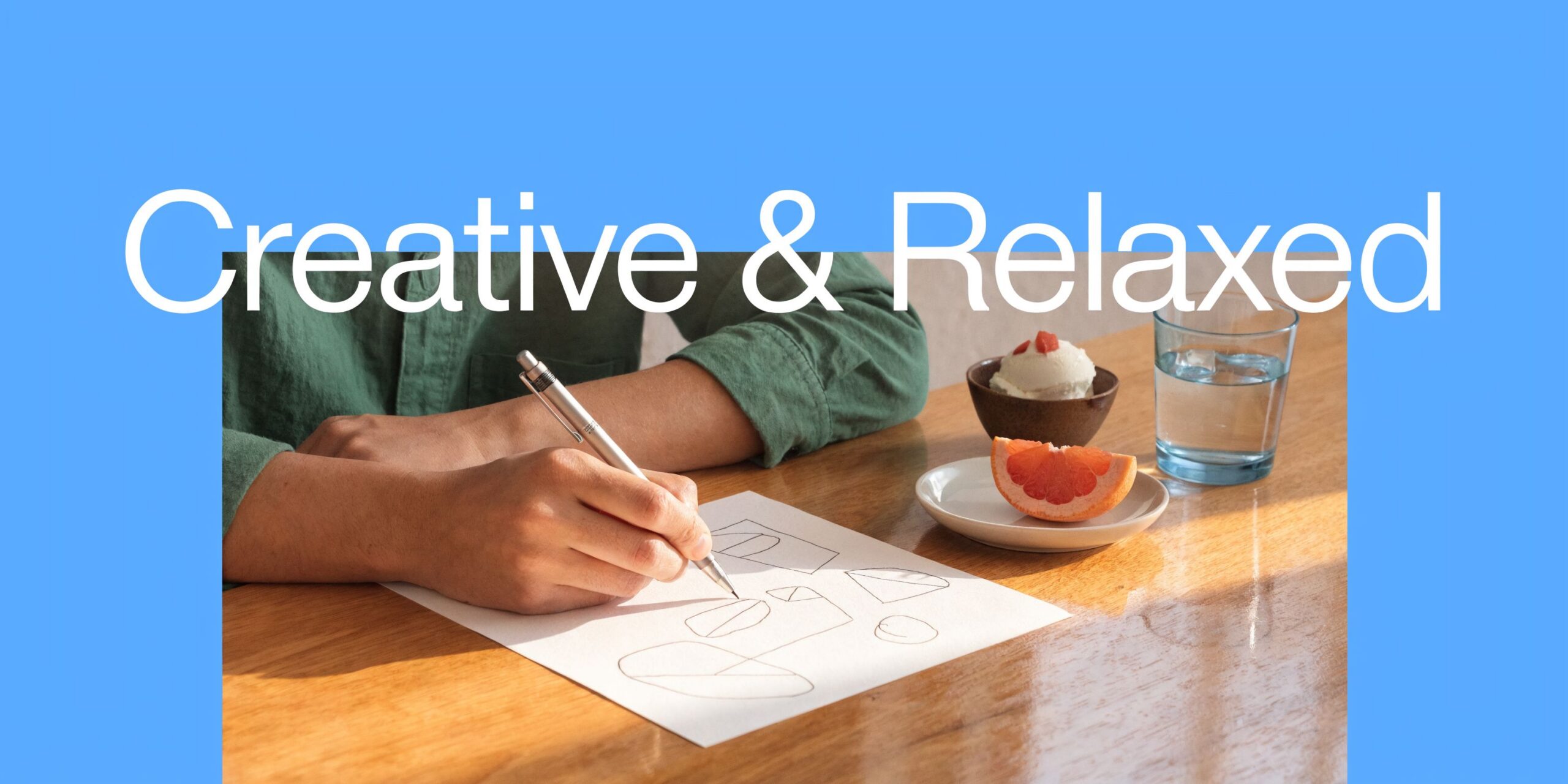 A person sitting at a wooden table sketching on paper near a serving of grapefruit and gelato.