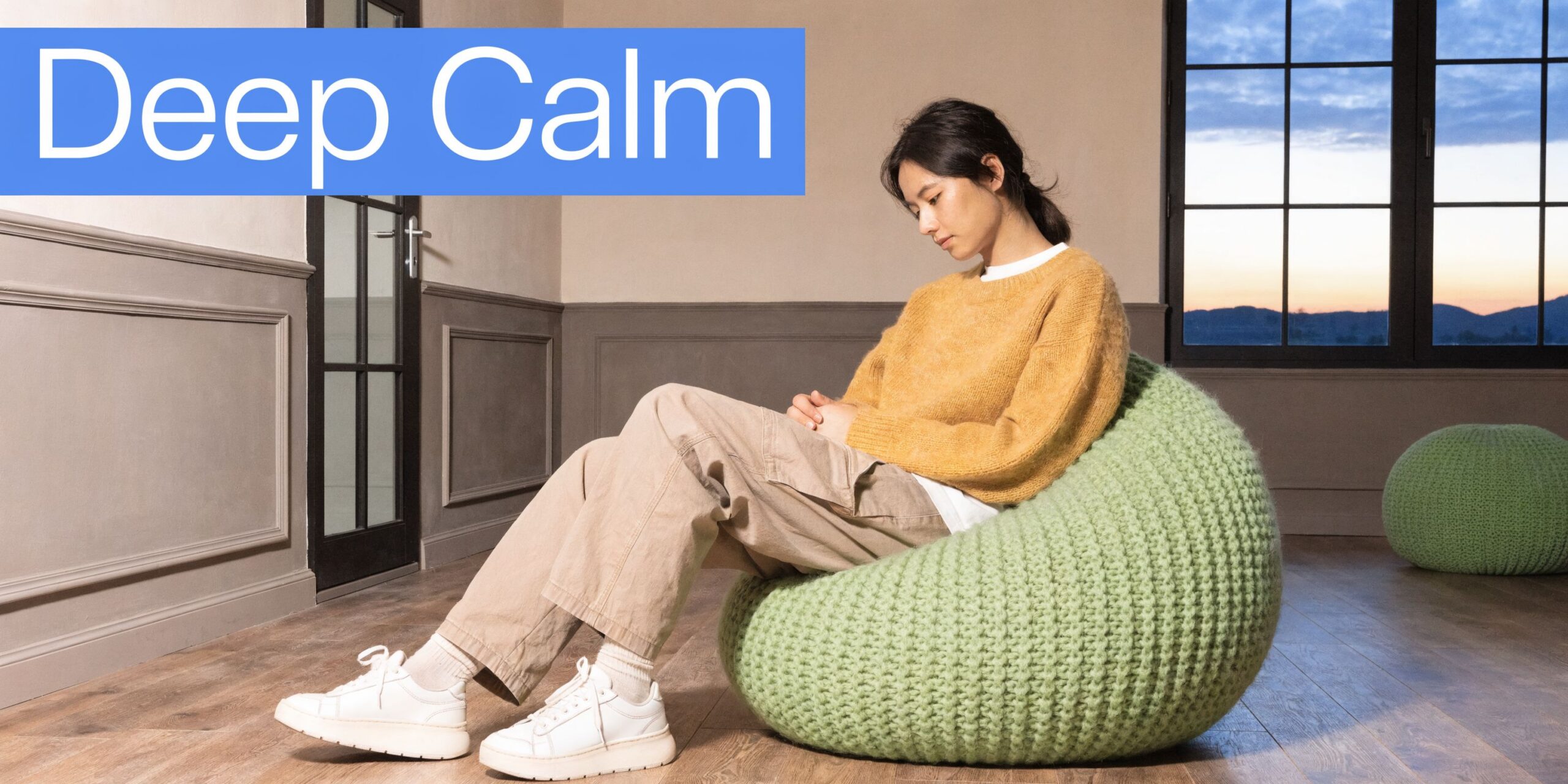 A person sitting peacefully on a large, knitted green bean bag chair in a cozy, minimalist room.