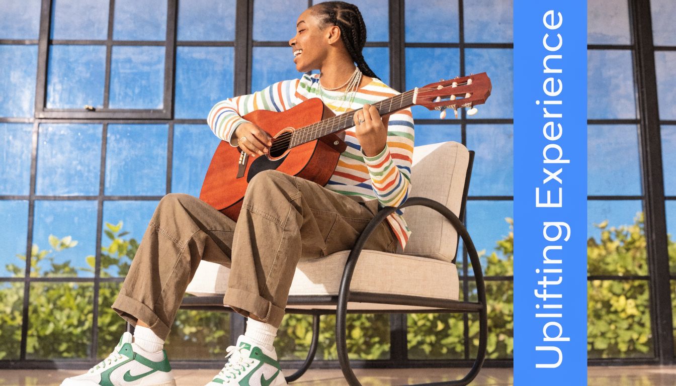 A happy person in a colorful striped sweater plays an acoustic guitar while sitting on a chair.