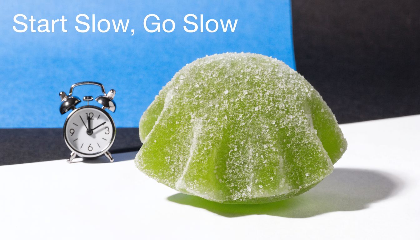 A green gummy candy sits next to a small silver alarm clock on a white surface.