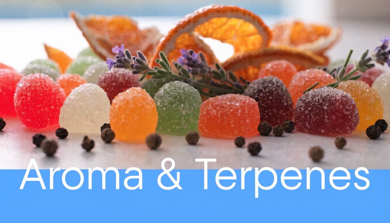 A close-up of colorful sugar-coated gummy candies, dried orange slices, lavender, and black peppercorns on white.